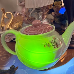 May include: A vibrant, lime green ceramic teapot with a matching handle and spout. The lid is a muted pink with a checkered pattern, and the body features floral accents. The teapot is a decorative item.
