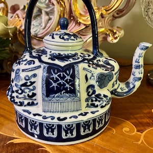 May include: A blue and white ceramic teapot with a curved spout and a large handle. The teapot features intricate patterns and designs, including floral and geometric motifs. The lid has a small blue knob. The teapot is placed on a wooden surface.