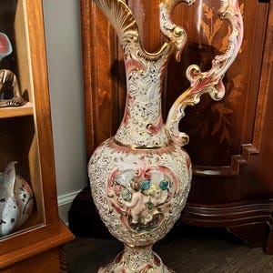 May include: A tall, ornate ceramic pitcher with a white base, adorned with pink and gold accents. The pitcher features a detailed handle and spout, with a central design of cherubs. The overall design is vintage and decorative.