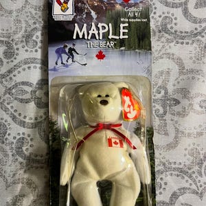 May include: A white plush bear toy named "Maple" from the Ty Beanie Baby collection, packaged in a clear plastic case. The bear has a red ribbon and a small Canadian flag. The packaging features a Canadian theme with hockey players and a mountain scene.