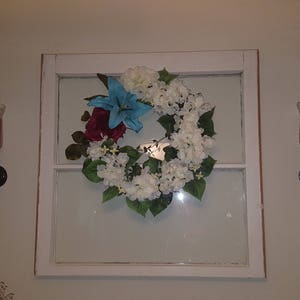 May include: A white window frame with a white and blue floral wreath hanging in the center. Two black candle holders with red candles are on either side of the window.