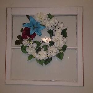 May include: A white painted window frame with a floral wreath inside. The wreath is made of white and blue flowers with green leaves.