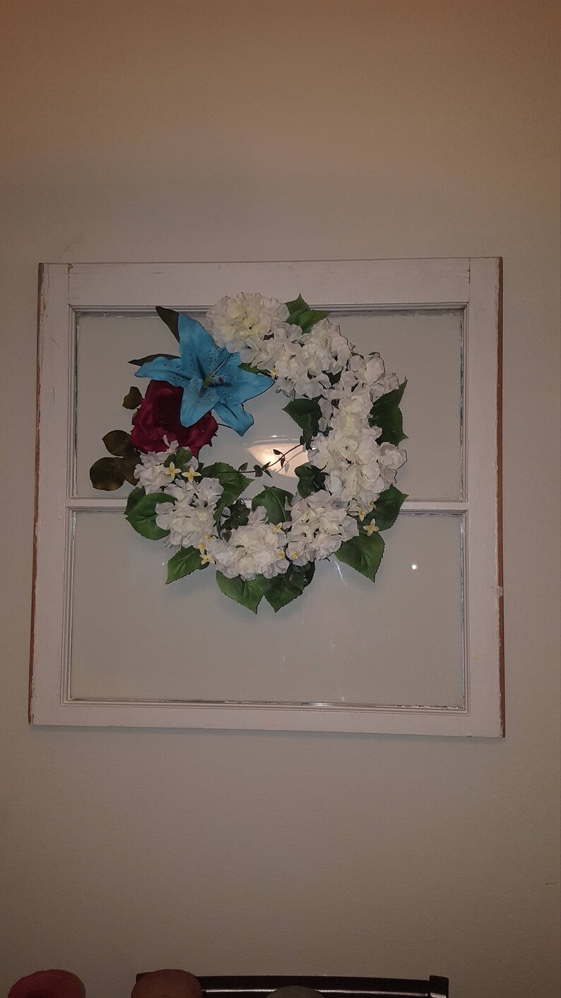 May include: A white painted window frame with a floral wreath inside. The wreath is made of white and blue flowers with green leaves.