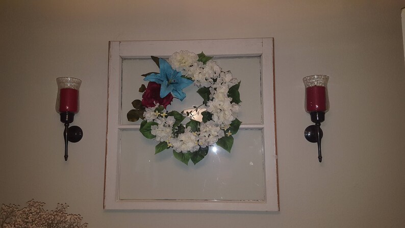 May include: A white window frame with a white and blue floral wreath hanging in the center. Two black candle holders with red candles are on either side of the window.