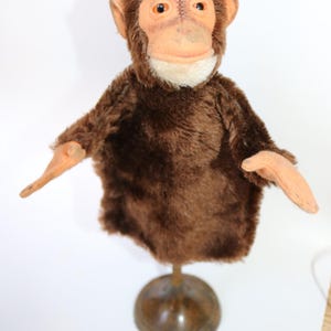 May include: A brown plush monkey puppet with orange eyes and a white mouth. The puppet is on a wooden stand.