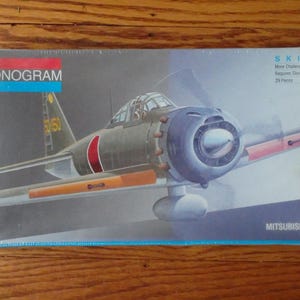 May include: A model kit box for a Mitsubishi Zero fighter plane. The box is blue and white with a picture of the plane on the front. The box says "Skill 2" and "Monogram" on the front. The box also says "1-48 Mitsubishi Zero" on the front.