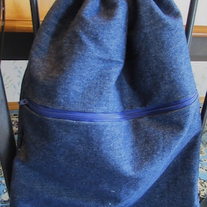 May include: A blue denim drawstring backpack with a zippered pocket. The top of the bag has a colorful floral pattern. The bag is sitting on a chair.