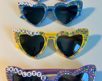 May include: Four pairs of heart-shaped sunglasses with rhinestones. Each pair has a different color frame and a different name spelled out in white letters on the top: Thea, Nikki, Lindsey, and Charlotte.