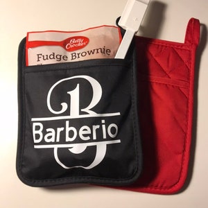 May include: Black and red oven mitts with a white monogram that says "JB Barberio". The mitts have a pocket on the front that holds a package of Betty Crocker Fudge Brownie mix and a white spatula.