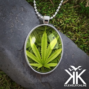 Puede incluir: Un collar de cadena de plata con un colgante redondo que presenta una hoja de marihuana verde encapsulada en vidrio.