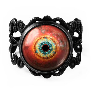May include: A black metal ring with an ornate design features a large, round, red eye with a yellow and blue iris and a black pupil.