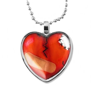 May include: A silver chain necklace with a heart-shaped pendant. The pendant is red and has a white bandage and stitches on it.