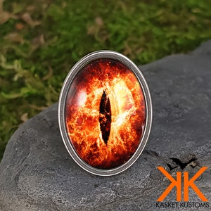 May include: A silver ring with a fiery orange and red eye design. The eye is surrounded by a silver ring and has a black pupil.