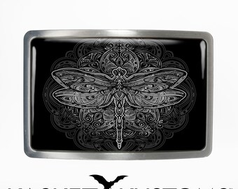 May include: A silver belt buckle with a black background featuring a white detailed dragonfly design. The dragonfly is surrounded by a circular mandala pattern.