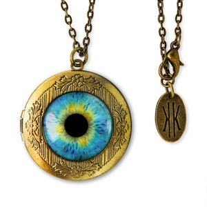 May include: A gold locket necklace with a blue eye design. The locket has a detailed floral pattern around the edge. The necklace has a small tag with the letters "JK" on it.
