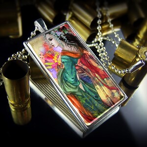 May include: A rectangular pendant with a silver frame and a detailed illustration of a woman in a colorful kimono. The pendant hangs on a silver chain. The background includes brass bullet casings.