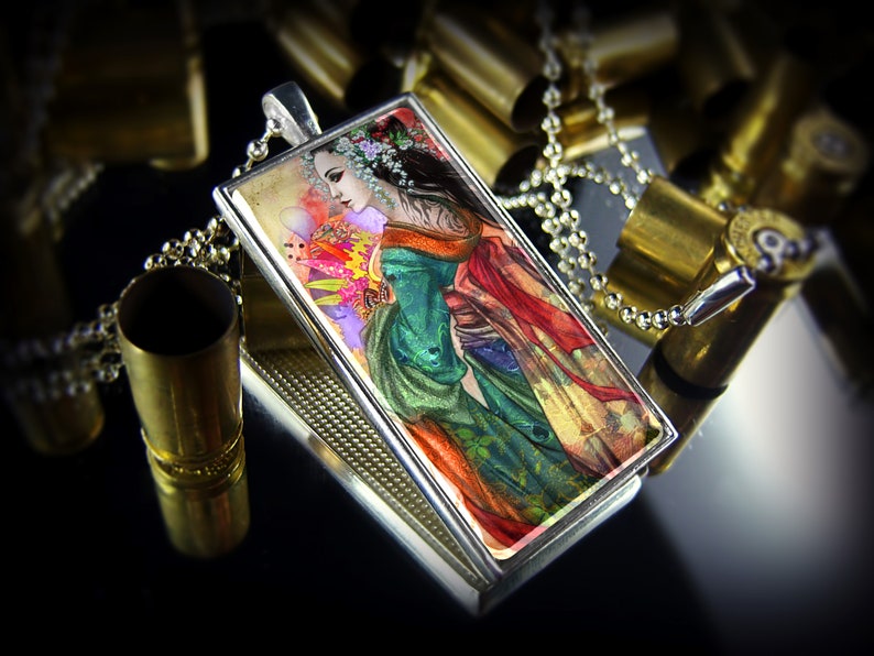 May include: A rectangular pendant with a silver frame and a detailed illustration of a woman in a colorful kimono. The pendant hangs on a silver chain. The background includes brass bullet casings.