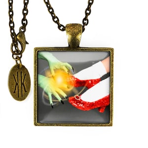 Puede incluir: Un collar con un colgante cuadrado y una cadena plateada. El colgante presenta una imagen colorida de una mano verde que alcanza un par de zapatos rojos.