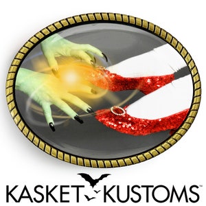 May include: A gold-toned belt buckle with a round, glass-like center featuring a green hand with black nails reaching towards a pair of red sequined shoes. The buckle is surrounded by a gold-toned, rope-like border. The text "KASKET KUSTOMS" is printed below the buckle.