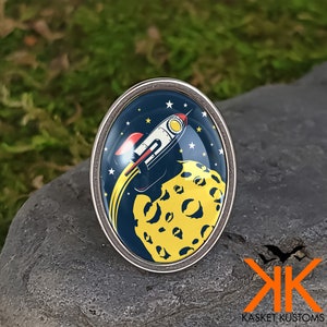 May include: A silver oval-shaped pin with a blue background and a yellow moon with craters. A red and white rocket is flying past the moon with a yellow trail.