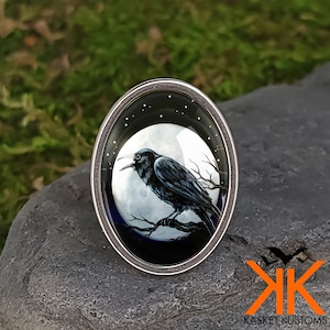 May include: A silver ring with a black raven perched on a branch in front of a full moon. The moon is surrounded by stars and the background is a dark blue.