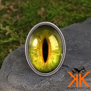 May include: A silver ring with a yellow and orange eye design. The eye is set in a glass cabochon.