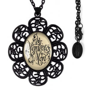 May include: Black necklace with a large, ornate flower pendant featuring a glass cabochon with the words "Memento Mori" in a decorative script. The pendant is attached to a black chain with a small black charm.