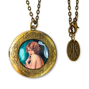 May include: A gold locket necklace with a vintage-style image of a woman with red hair and a blue and green background. The locket has a detailed floral design around the edge. The necklace has a simple chain and a small oval tag with the initials "JK" on it.
