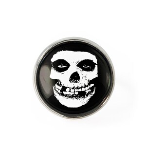 Könnte beinhalten: Ein silberfarbener Knopf mit schwarzem Hintergrund und einem weißen Misfits-Schädel-Logo. Der Schädel hat ein breites Grinsen und zwei große Augenhöhlen.