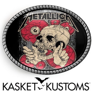 Puede incluir: Una hebilla de cintur&oacute;n de metal negro y rojo con un dise&ntilde;o de calavera y huesos cruzados. La hebilla tiene la palabra "Metallica" en letras blancas.