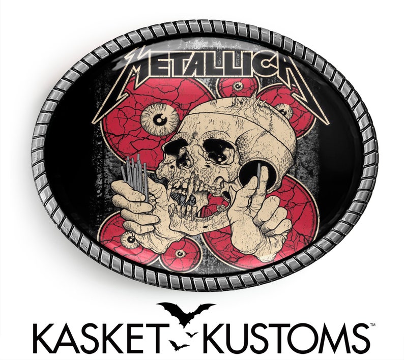Puede incluir: Una hebilla de cintur&oacute;n de metal negro y rojo con un dise&ntilde;o de calavera y huesos cruzados. La hebilla tiene la palabra "Metallica" en letras blancas.
