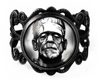 May include: A black metal ring with a black and white image of Frankenstein's monster. The ring has an ornate filigree design around the central image. The ring is a gothic or Halloween-themed accessory.