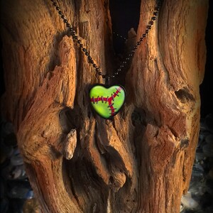 Pode incluir: Um pingente verde em forma de cora&ccedil;&atilde;o com pontos vermelhos pendurado em uma corrente preta. O pingente est&aacute; em um peda&ccedil;o de madeira &agrave; deriva.