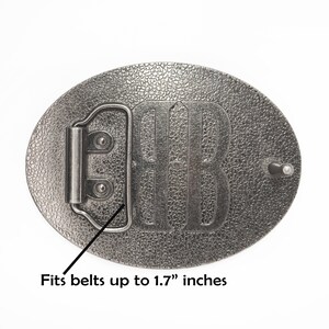 May include: Silver-toned metal belt buckle with a textured surface. The buckle features a raised design of the letter "B". It fits belts up to 1.7 inches wide.