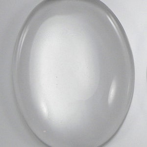 May include: A clear glass cabochon, oval shaped, with a smooth, polished surface.