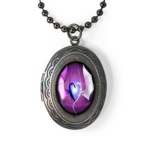Puede incluir: Un collar con un medallón plateado con una imagen de dos dragones entrelazados con un corazón en el centro, sobre un fondo morado y blanco.