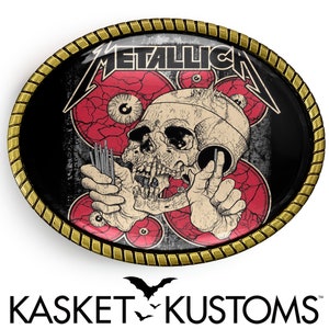 Puede incluir: Una hebilla de cintur&oacute;n ovalada negra y dorada con un dise&ntilde;o de calavera y huesos cruzados. La calavera sostiene un juego de agujas y est&aacute; rodeada de ojos rojos. El texto "METALLICA" est&aacute; impreso en blanco en la hebilla.