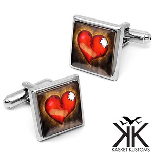 May include: A pair of silver cufflinks with a square design. Each cufflink features a red heart with a bandage on it, set against a dark brown background.