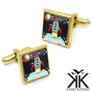 May include: Gold-toned square cufflinks featuring a retro rocket ship design against a black background with a planet and stars. The rocket is red, blue, and yellow, with a light blue cloud base. The cufflinks have a shiny finish.