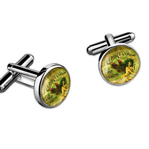Könnte beinhalten: Ein Paar silberne Manschettenknöpfe mit einem runden, grün-goldenen Design, das eine Fee mit Flügeln und den Text "La Fée Verte" zeigt.