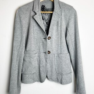 Op de afbeelding: Een grijze blazer met twee knopen en twee zakken. De blazer heeft een zwarte en witte bloemenprint voering.