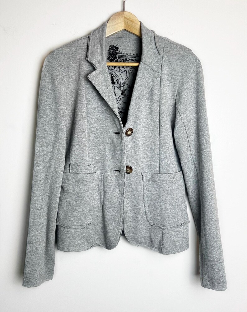 Op de afbeelding: Een grijze blazer met twee knopen en twee zakken. De blazer heeft een zwarte en witte bloemenprint voering.