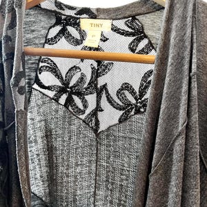 以下が含まれることがあります： グレーのカーディガンに黒い花柄のレースを重ねたもの。カーディガンはフード付きで、木製ハンガーにかけられています。カーディガンのタグには「TINY」と書いてあります。