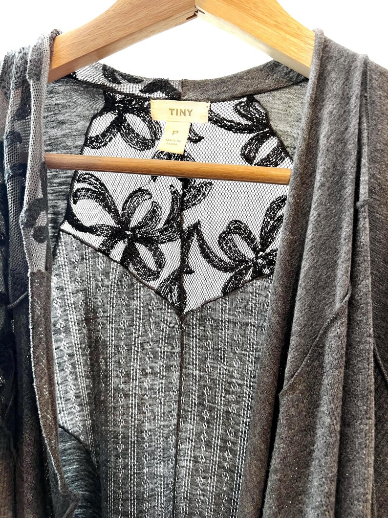 以下が含まれることがあります： グレーのカーディガンに黒い花柄のレースを重ねたもの。カーディガンはフード付きで、木製ハンガーにかけられています。カーディガンのタグには「TINY」と書いてあります。
