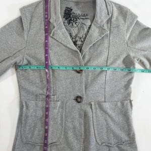 Op de afbeelding: Grijze blazer met twee zakken en een knoopsluiting. De blazer heeft een gedrukt label aan de binnenkant van de kraag met de tekst "Paris is a state of mind".