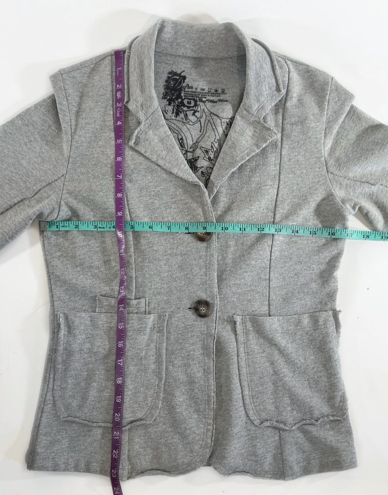 Op de afbeelding: Grijze blazer met twee zakken en een knoopsluiting. De blazer heeft een gedrukt label aan de binnenkant van de kraag met de tekst "Paris is a state of mind".