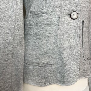 Op de afbeelding: Een grijze blazer met lange mouwen, een enkele knoopsluiting en een klepzak aan de linkerkant.