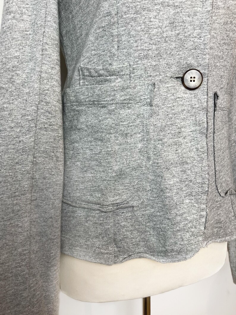 Op de afbeelding: Een grijze blazer met lange mouwen, een enkele knoopsluiting en een klepzak aan de linkerkant.