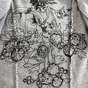 Op de afbeelding: Een grijs T-shirt met een zwart bloemenontwerp. Het ontwerp heeft een grote bloem met een cirkel in het midden en kleinere bloemen en bladeren eromheen. De tekst "River" staat op het label.