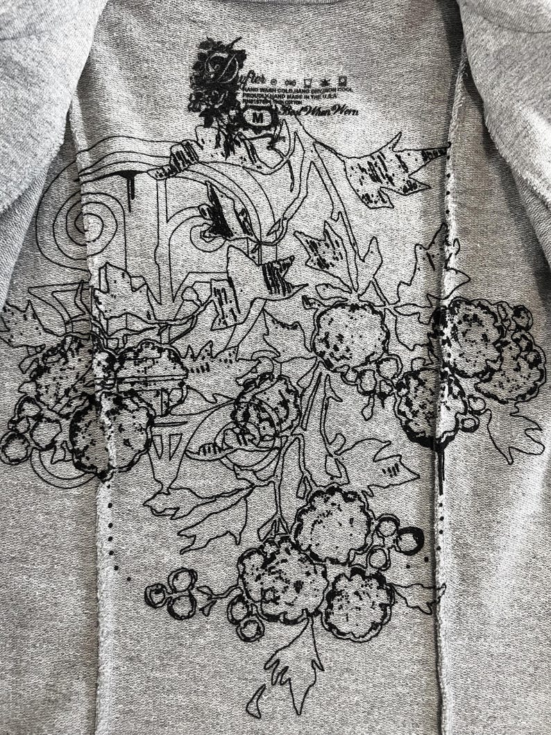 Op de afbeelding: Een grijs T-shirt met een zwart bloemenontwerp. Het ontwerp heeft een grote bloem met een cirkel in het midden en kleinere bloemen en bladeren eromheen. De tekst "River" staat op het label.